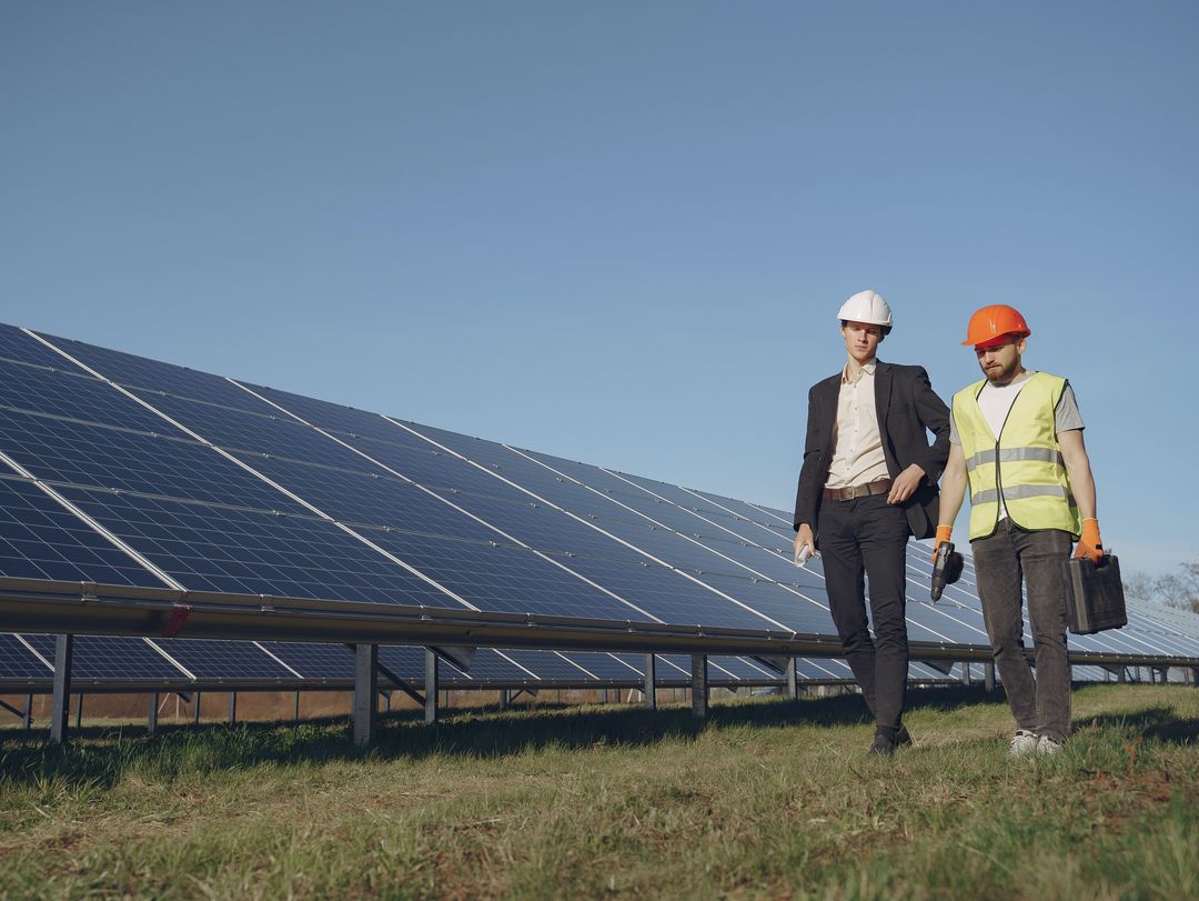 Stockfoto. Ein Mann in Anzug und ein Mann mit Warnweste und Werkzeugkiste laufen vor einer Solaranlage her.